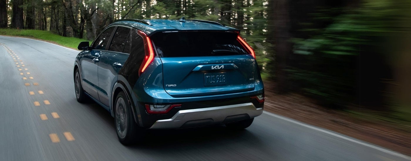 A blue 2024 Kia Niro PHEV is shown from the rear driving on a tree-lined road.